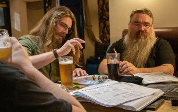 Two individuals, one with long light brown hair and a medium beard, the other with short grey hair and a long grey beard, both with glasses, sit around a pub table, which has drinks, character sheets, source books and other RPG material on. The person on the left has just rolled dice.